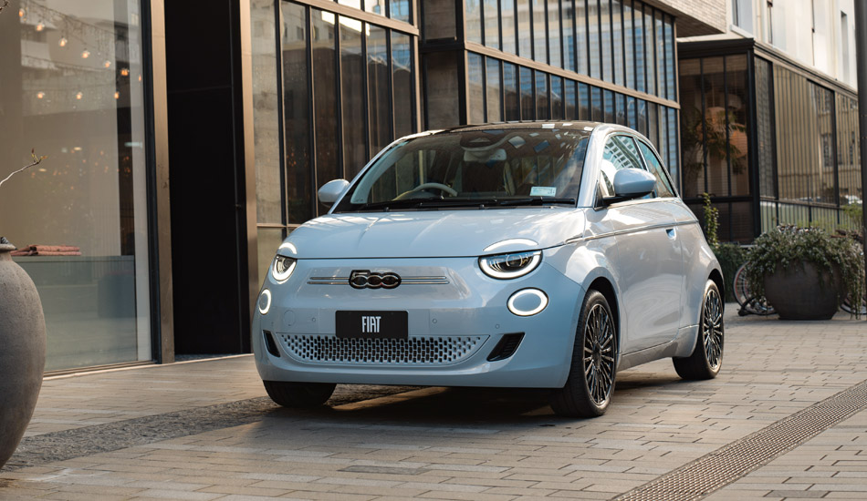 A silver Fiat 500e electric hatchback parked on a cobblestone street in front of a modern building